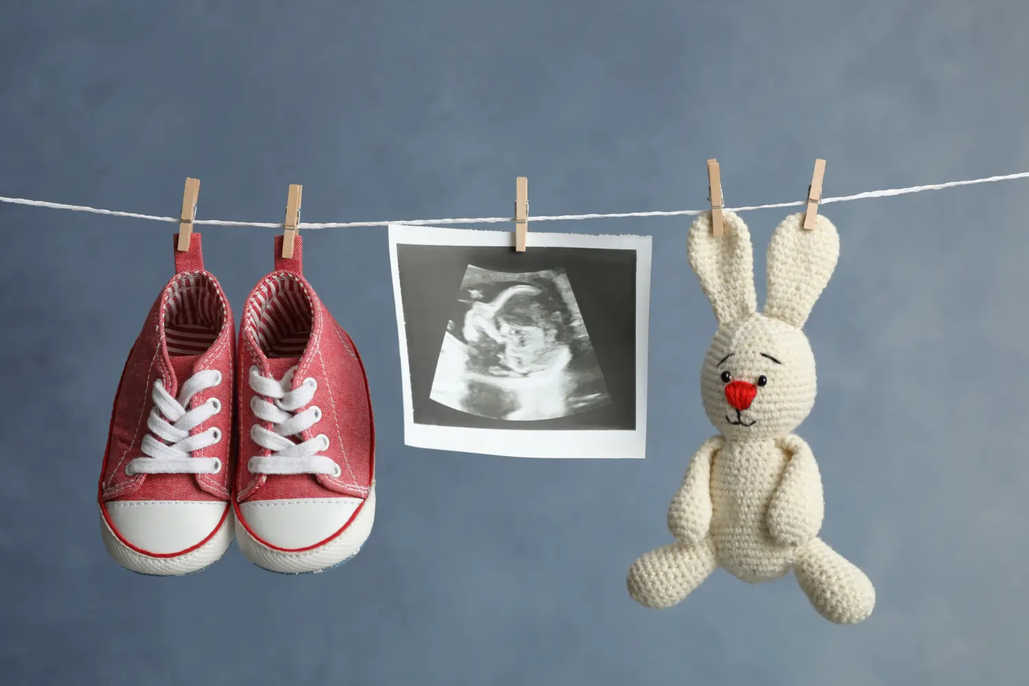 Paar Kinderschuhe, Ultraschallfoto und Spielzeughase hängen an der Wäscheleine vor dunklem Hintergrund